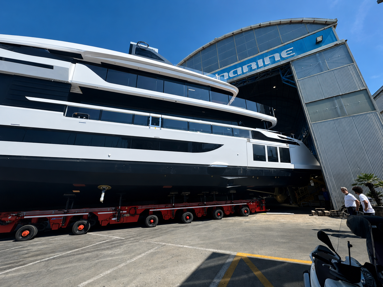 Large white yacht outside a shipyard building on a trailer, with people nearby